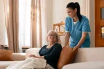 Caregiver in scrubs assisting elderly woman on couch