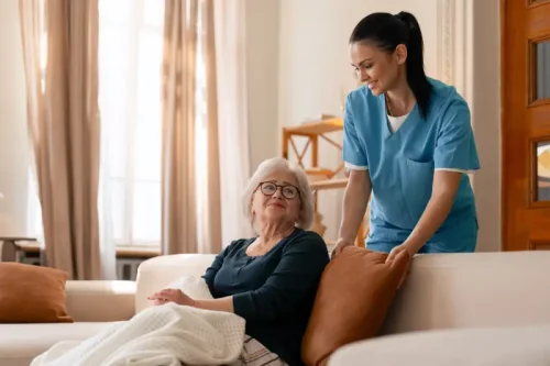 Caregiver in scrubs assisting elderly woman on couch