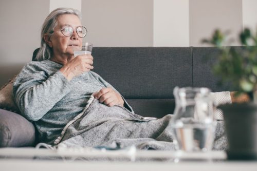 Elderly woman in glasses sits on a gray sofa, wrapped in a blanket, drinking water. A peaceful ambiance with a blurred plant in the foreground.