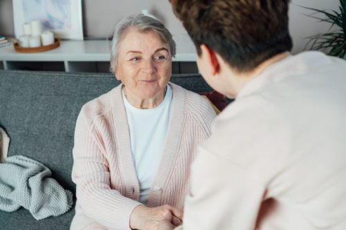 An elderly woman in a pink cardigan smiles warmly at a person holding her hand on a sofa. The scene conveys comfort and connection.