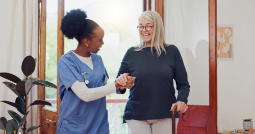 A caregiver in blue scrubs assists a smiling elderly woman using a cane. They share a warm moment in a bright, cozy living room with natural light.