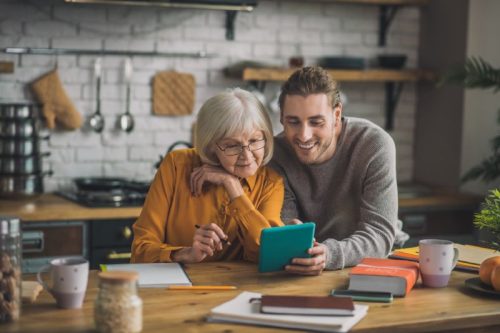 An elderly woman and a young man sit at a kitchen table, smiling at a tablet. The cozy kitchen includes books, mugs, and a plant, conveying warmth and connection.