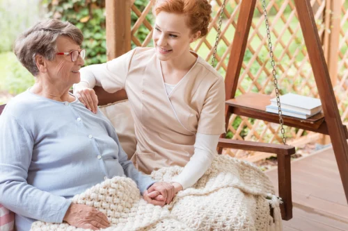 A young caregiver in a beige uniform sits beside an elderly woman on a porch swing. They smile warmly, holding hands under a knitted blanket.